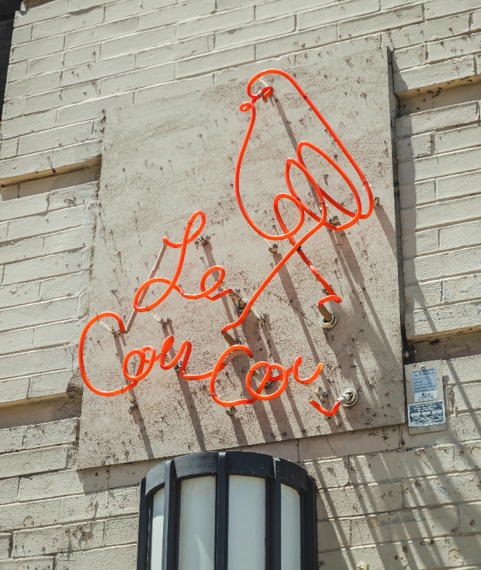 A red neon sign reading "Le Cou Cou" with a bird illustration mounted on a brick exterior wall.