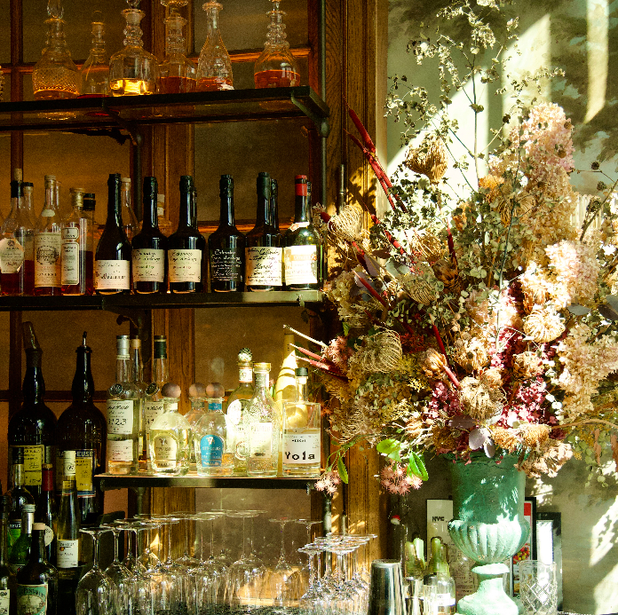 Vintage bar shelves with wine bottles and decanters next to a large dried flower arrangement in green vase by sunny window