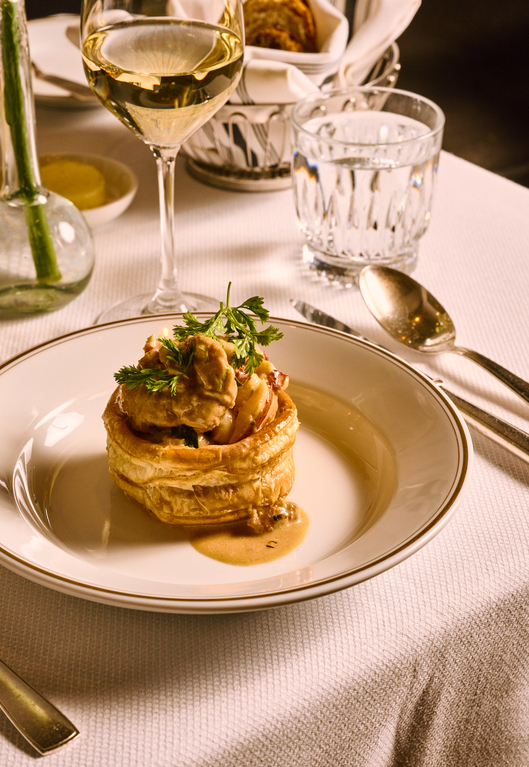 Gourmet dish in a pastry basket, topped with herbs, served with white wine on a white plate with cutlery.