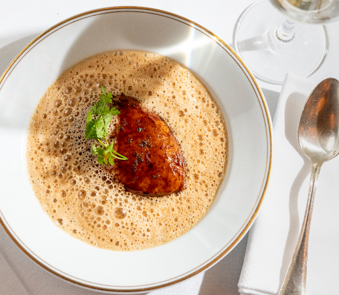 A seared protein with herb garnish in a frothy cream sauce in a gold-rimmed bowl, served with a spoon and wine glass.