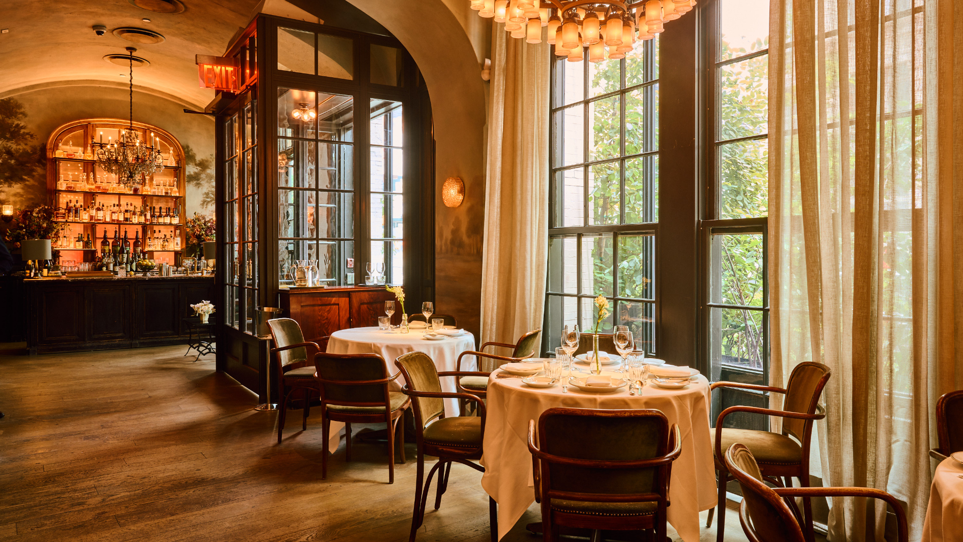 A cozy restaurant interior with vintage decor, wooden floors, floral wallpaper, a bar with arched shelves and chandelier, and a round table set for two.