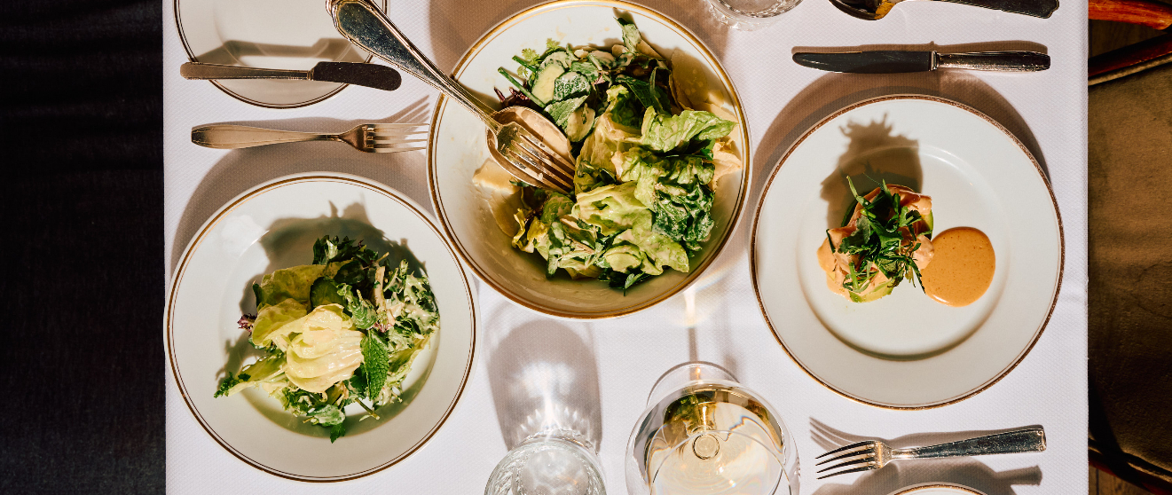 An overhead view of three plated dishes with green salads and sauces on a white tablecloth with wine glasses and silverware.
